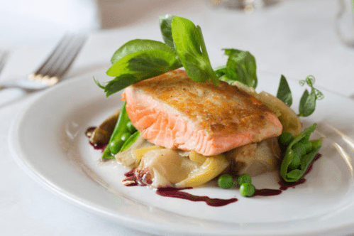 A plated dish featuring grilled salmon on a bed of vegetables, garnished with greens and a drizzle of sauce.